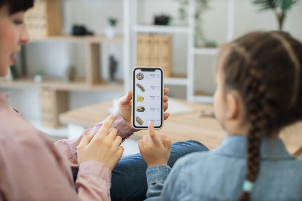 Back view of brunette woman and girl with french braids sitting at home and purchasing fast food online. Happy mother and daughter spending time together while using mobile phone for e-commerce.
