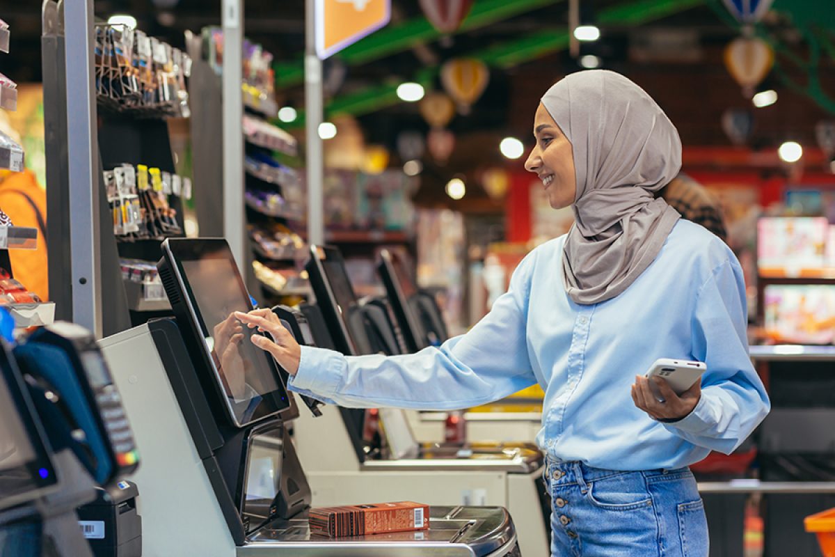 A woman buyer in a supermarket in a hijab pays for goods at a self-service checkout, convenient service for customers.